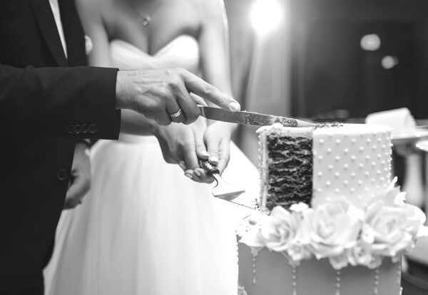 A bride and groom cut a tiered wedding cake together, holding the knife and server, with white roses decorating the cake.