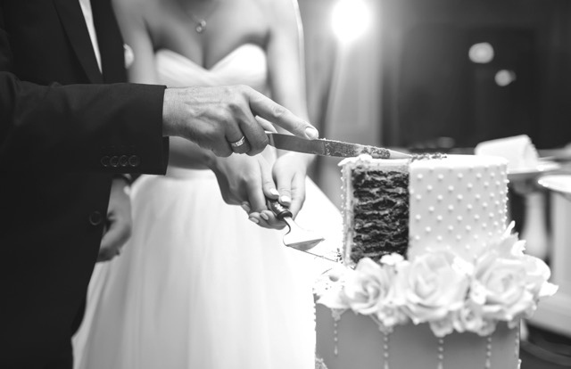 A bride and groom cut a tiered wedding cake together, holding the knife and server, with white roses decorating the cake.
