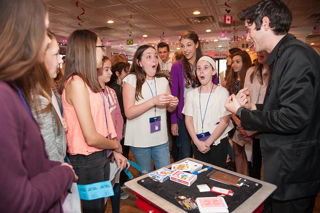 A magician performs a card trick for a group of excited kids and teens at a party, with surprised expressions and cards on a table.