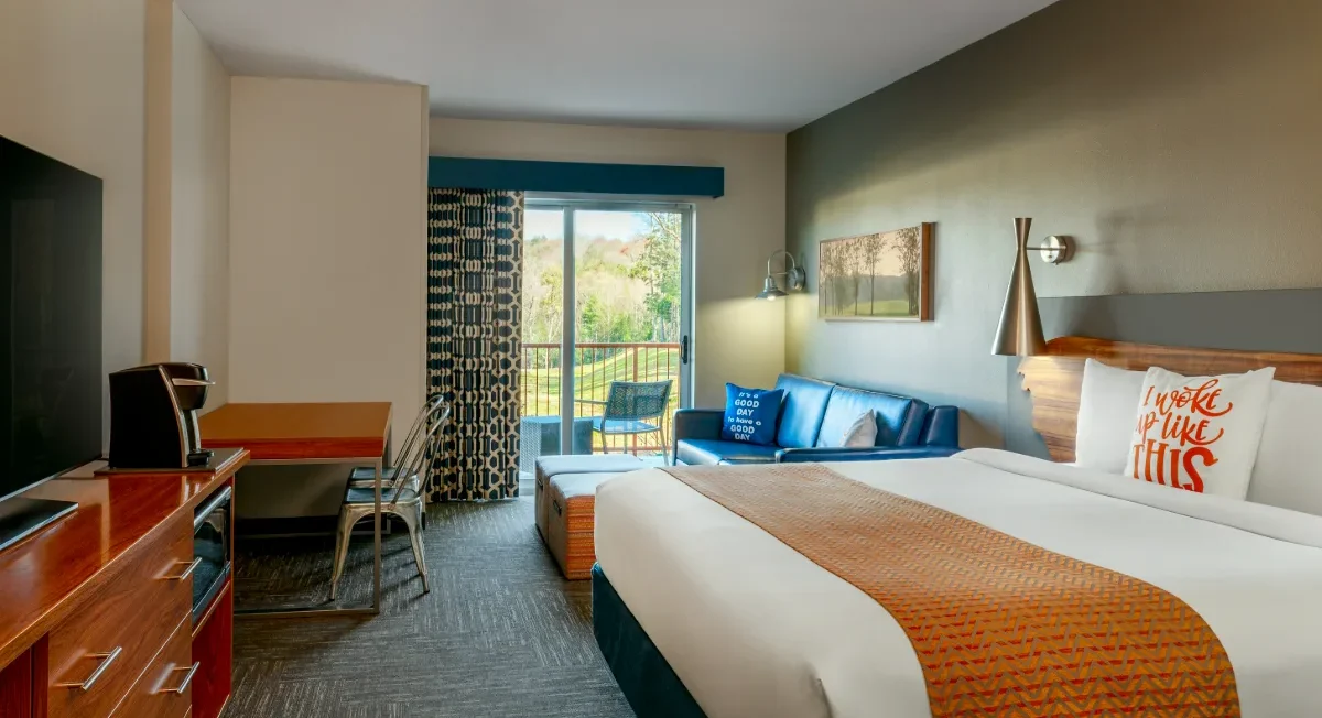 Modern hotel room with a king bed, desk, TV, blue sofa, and balcony with outdoor seating overlooking greenery.