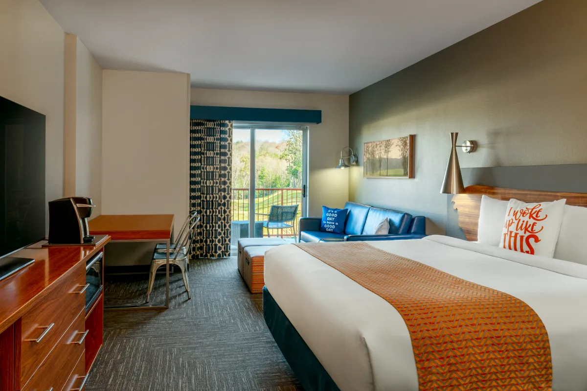 Modern hotel room with a king bed, desk, TV, blue sofa, and balcony with outdoor seating overlooking greenery.