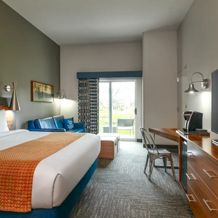 Modern hotel room with a king bed, blue sofa, desk, TV, and a sliding door opening to a patio with chairs outside.