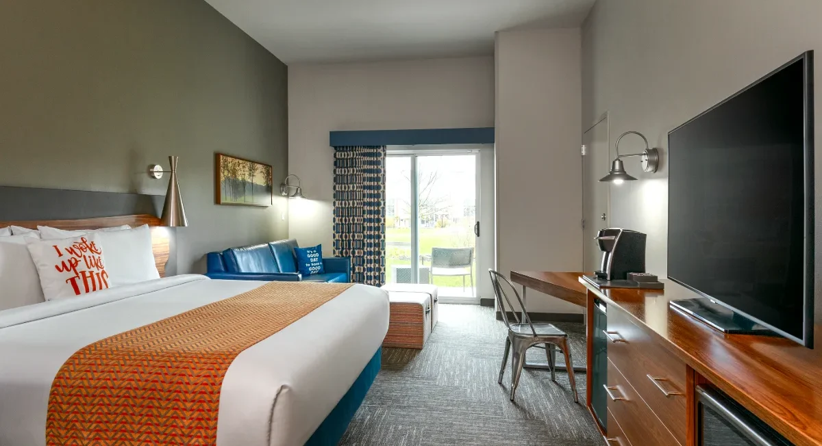 Modern hotel room with a king bed, TV, desk, blue sofa, sliding glass door, and patterned decor. Outdoor scenery visible through the door.