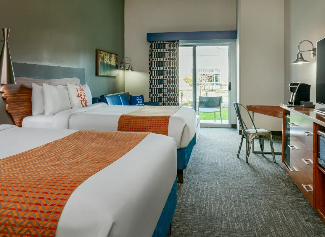 Modern hotel room with two beds, orange accents, a desk, TV, blue sofa, and sliding glass door leading to a patio with outdoor seating.