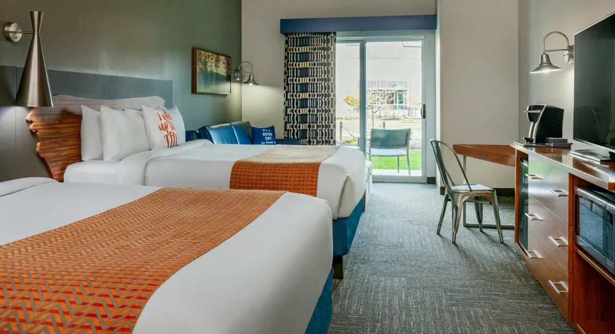 Modern hotel room with two beds, orange accents, a desk, TV, blue sofa, and sliding glass door leading to a patio with outdoor seating.