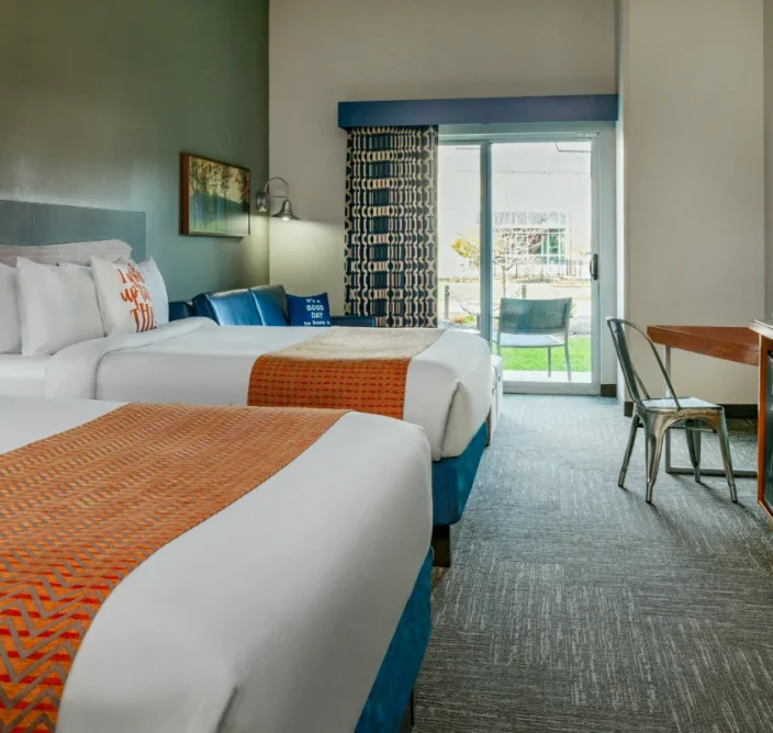 Modern hotel room with two beds, orange accents, a desk, TV, blue sofa, and sliding glass door leading to a patio with outdoor seating.
