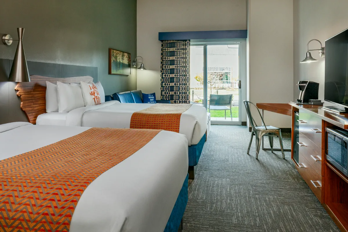 Modern hotel room with two beds, orange accents, a desk, TV, blue sofa, and sliding glass door leading to a patio with outdoor seating.