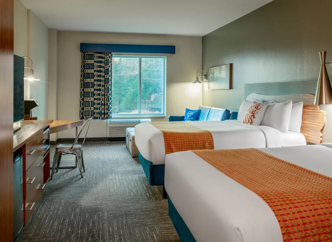 Hotel room with two double beds, orange blankets, a desk, chair, TV, armchair, window with trees outside, and modern decor.