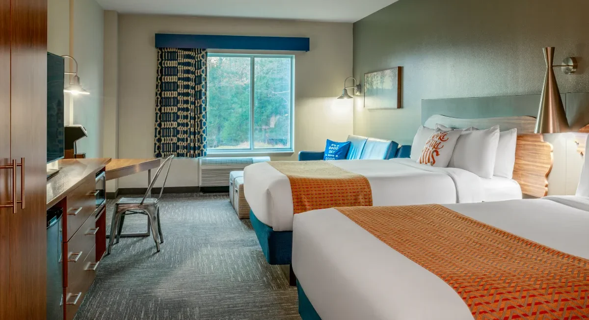 Hotel room with two double beds, orange blankets, a desk, chair, TV, armchair, window with trees outside, and modern decor.