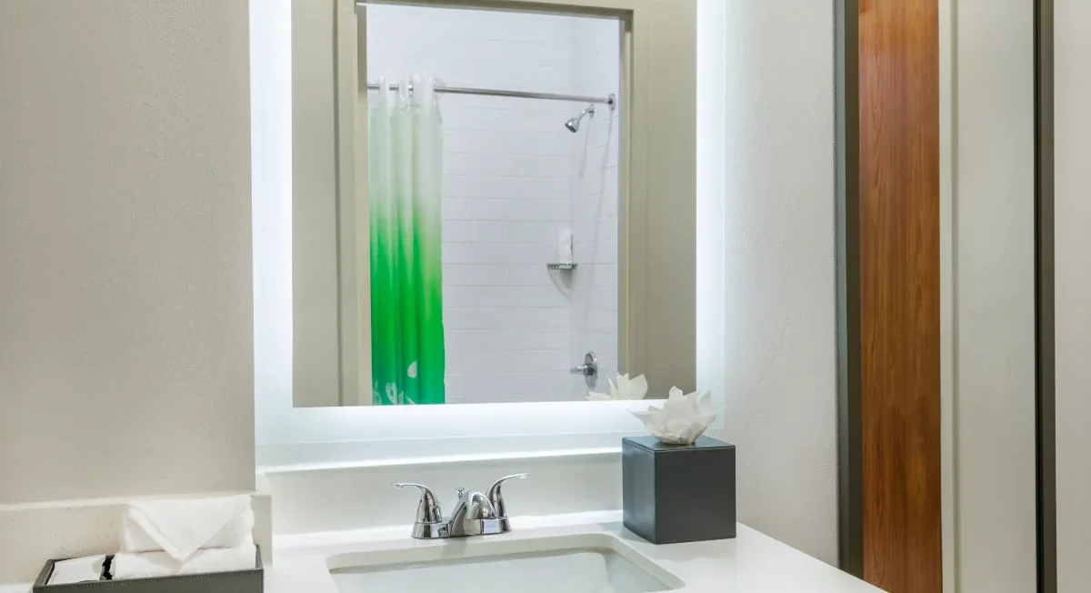 Modern bathroom with a sink, faucet, tissue box, towel tray, lighted mirror, and a shower with a green ombre curtain reflected in the mirror.