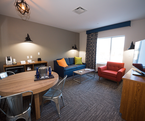 Modern hotel room with a round table, metal chairs, blue sofa, red armchair, TV, window with curtains, and a kitchenette area.