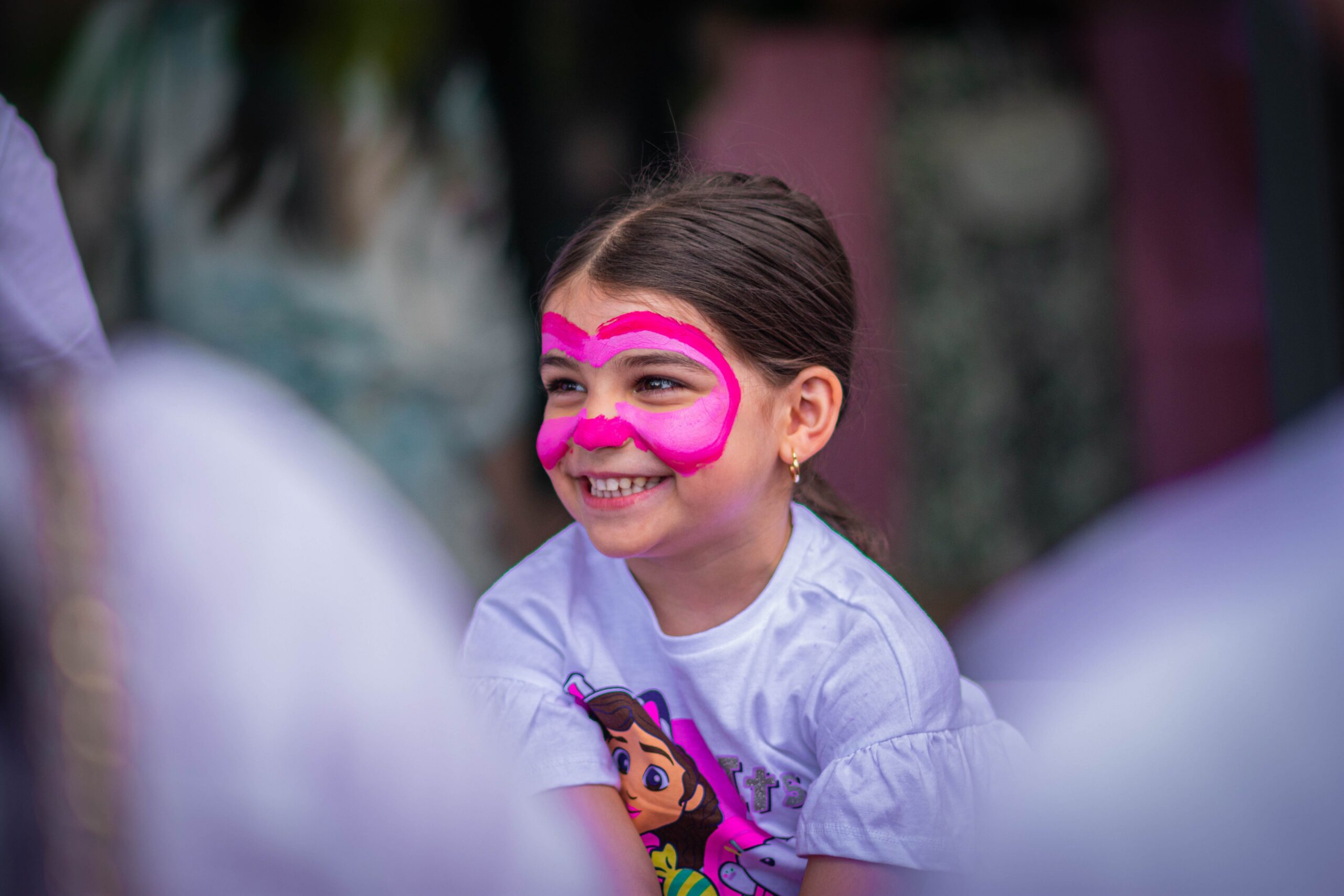 Smiling young girl with bright pink face paint and a cartoon character shirt, surrounded by blurred people in the foreground.