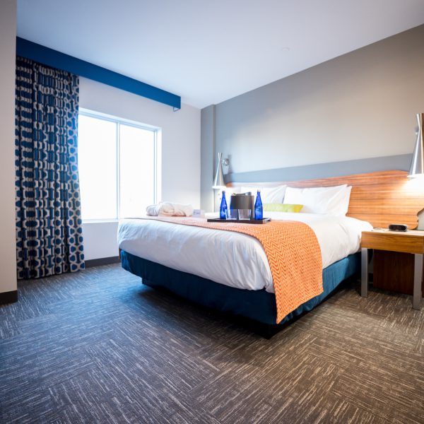 Modern hotel room with a large bed, wooden furniture, patterned curtains, and a tray with blue bottles on the bed near a bright window.