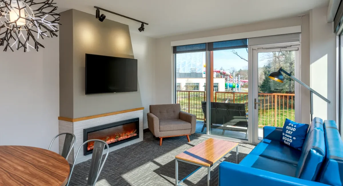 Modern living room with blue sofa, wall-mounted TV, electric fireplace, armchair, large window, and sliding door leading to a balcony with outdoor views.