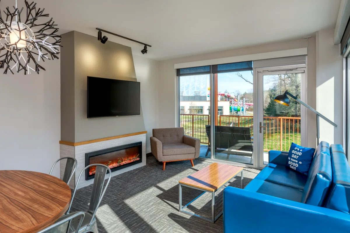 Modern living room with blue sofa, wall-mounted TV, electric fireplace, armchair, large window, and sliding door leading to a balcony with outdoor views.