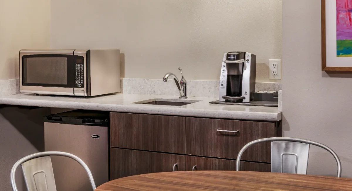 Small hotel kitchenette with microwave, mini fridge, sink, and coffee maker next to a round wooden table and metal chairs.