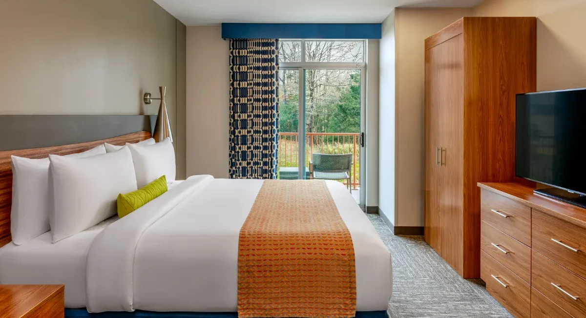 Modern hotel room with a neatly made bed, orange blanket, wardrobe, dresser, TV, and a window view of trees and a balcony.