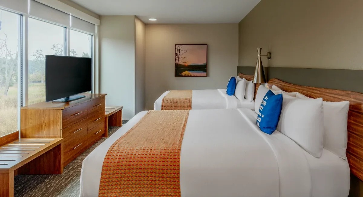 Hotel room with two double beds, orange throws, a TV on a dresser, bench, window with a view, and a framed landscape picture on the wall.