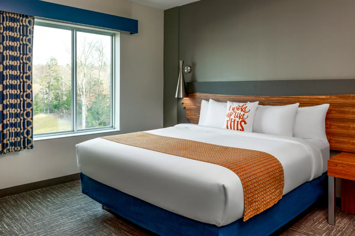 Modern hotel room with a large bed, orange runner, decorative pillow, window with trees outside, patterned curtains, and gray accent wall.