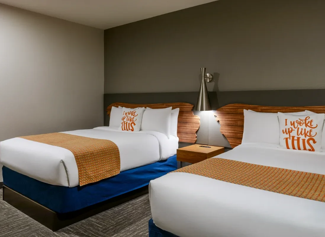 Hotel room with two beds, orange accent throws, pillows reading I woke up like THIS, a nightstand, lamp, and modern decor.