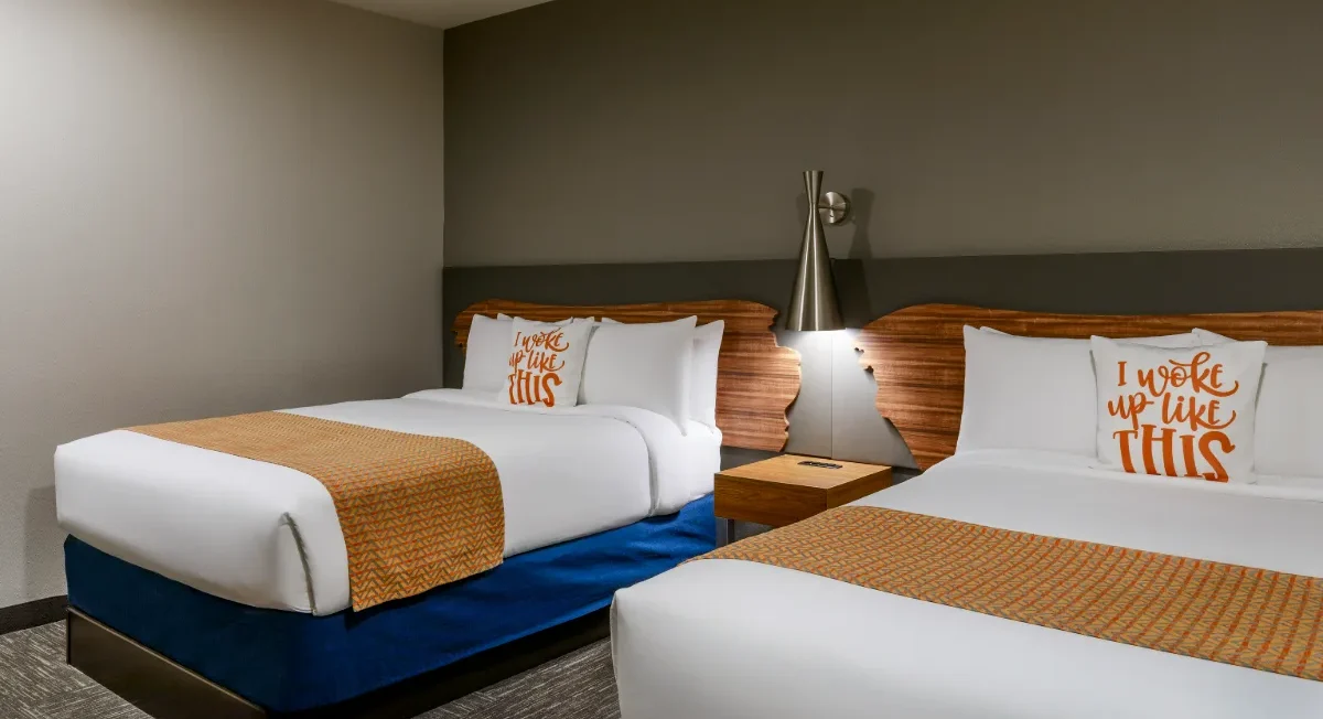Hotel room with two beds, orange accent throws, pillows reading I woke up like THIS, a nightstand, lamp, and modern decor.