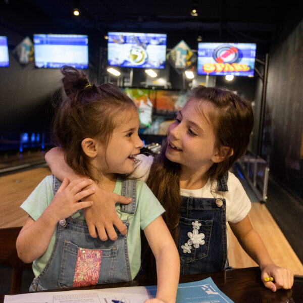 Two young girls in denim overalls smile and hug while coloring at a table in a bowling alley, with TV screens and bowling lanes in the background.