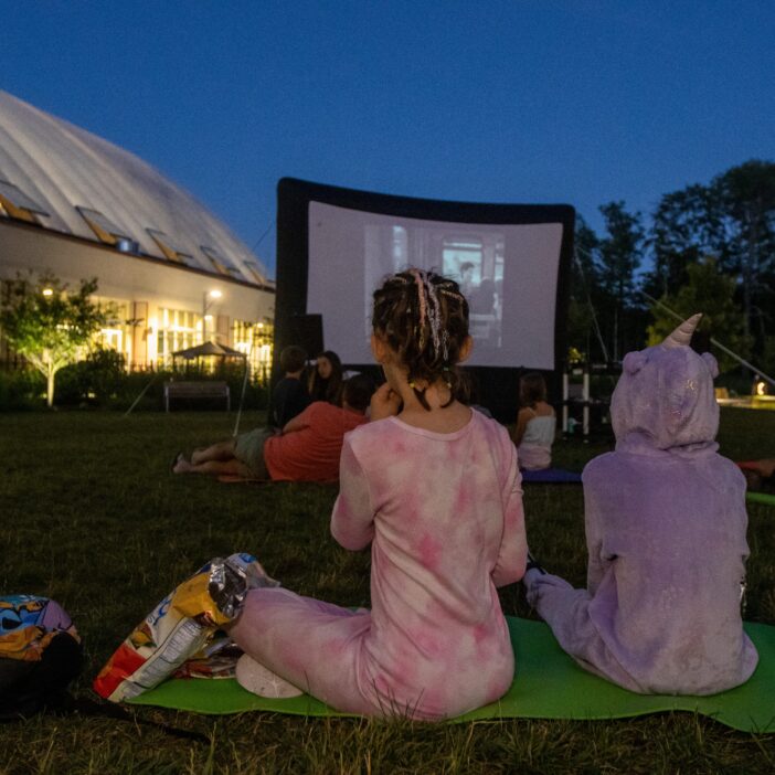 Children in pajamas watch an outdoor movie at dusk, sitting on mats on the grass in front of a large screen near a building.
