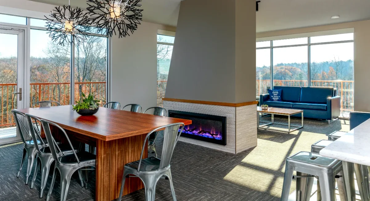 Modern dining and lounge area with a wooden table, metal chairs, fireplace, sofas, large windows, and views of trees outside.