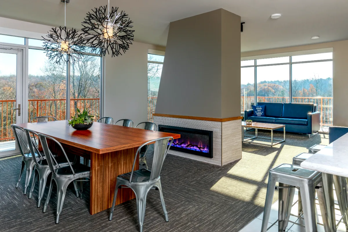 Modern dining and lounge area with a wooden table, metal chairs, fireplace, sofas, large windows, and views of trees outside.