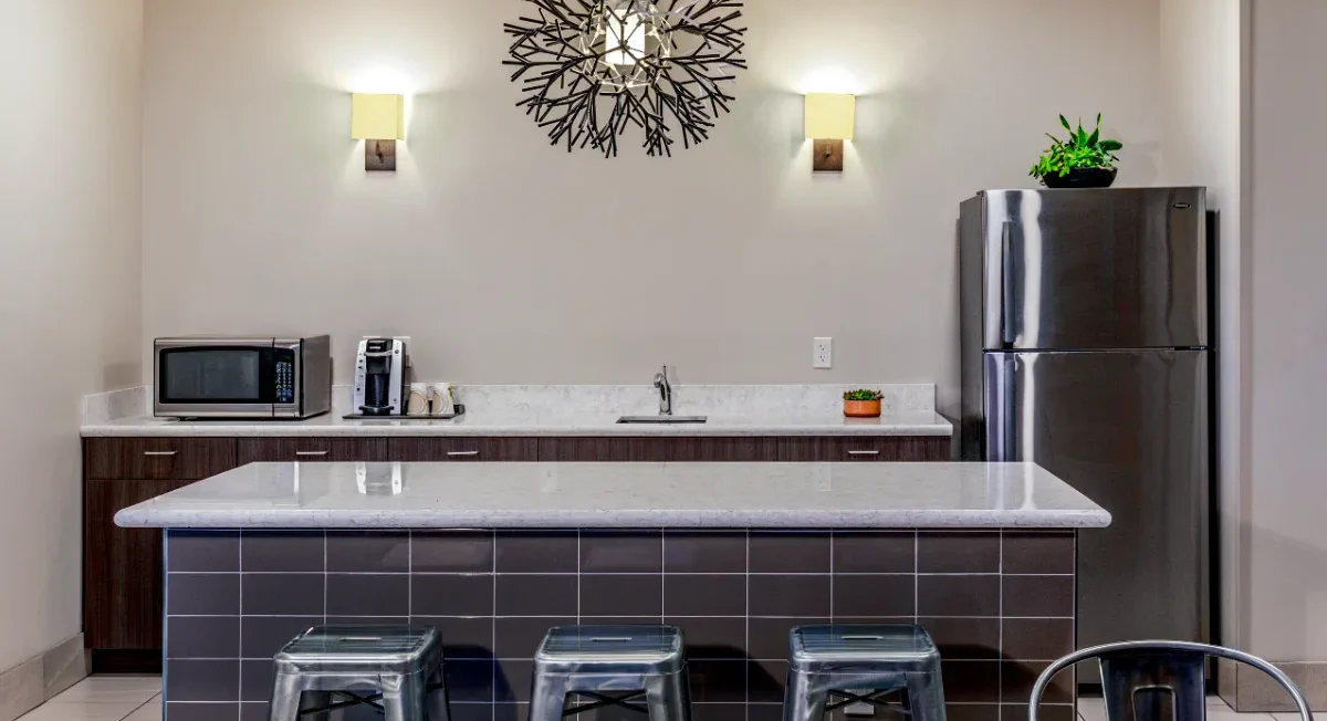 Modern kitchen with gray countertops, stainless steel fridge, microwave, coffee maker, and three clear barstools at a tiled island under decorative lighting.
