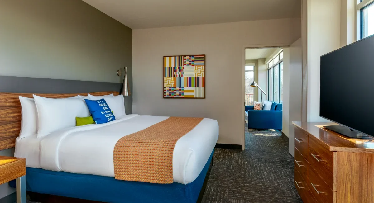 Modern hotel room with a king bed, colorful pillows, TV, dresser, artwork, and a view into a bright sitting area through an open doorway.
