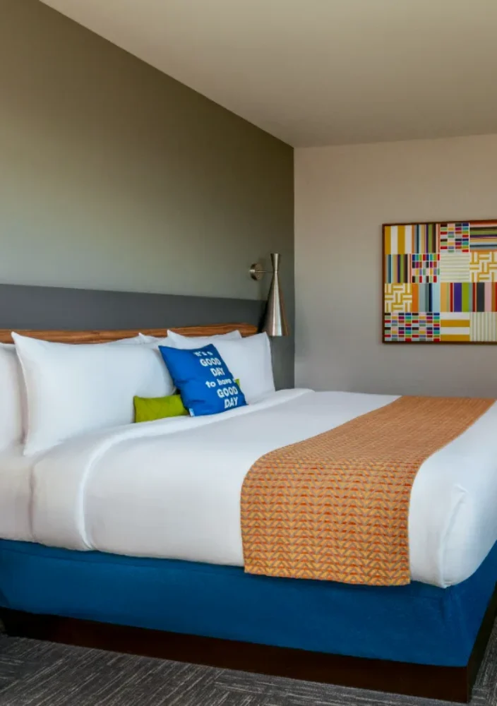 Modern hotel room with a king bed, colorful pillows, TV, dresser, artwork, and a view into a bright sitting area through an open doorway.