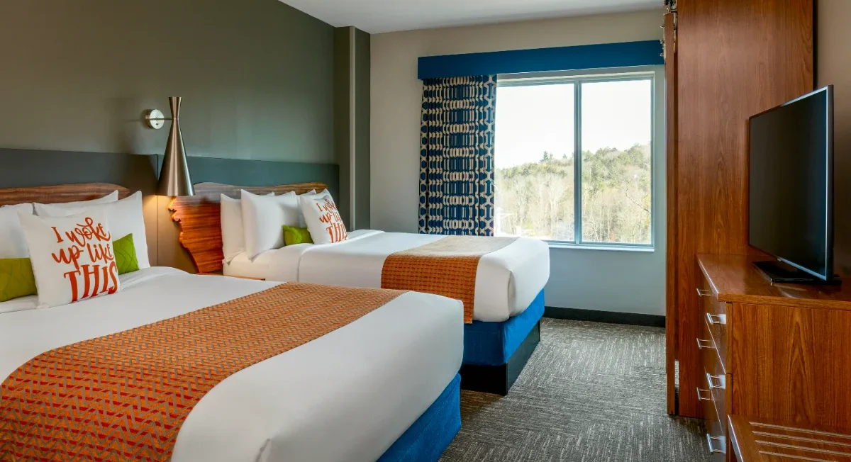 Hotel room with two double beds, orange accents, a flat-screen TV, large window with blue curtains, and modern decor.