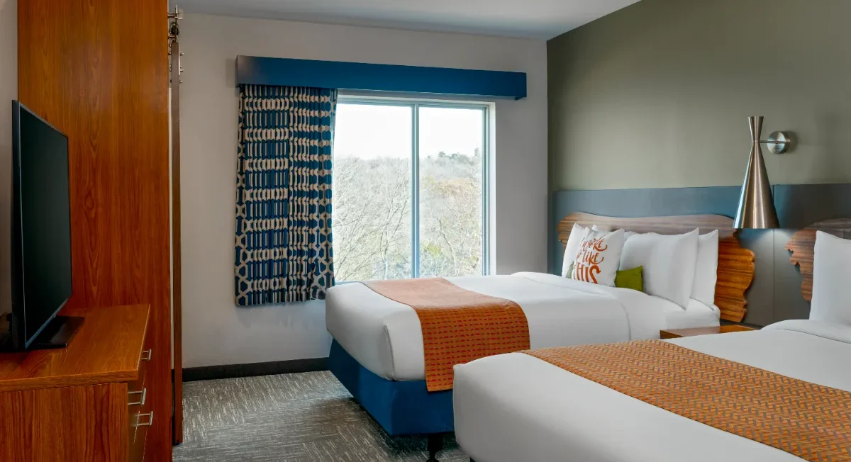 Hotel room with two beds, orange accents, a TV, wood furniture, modern lighting, and a window with patterned curtains showing trees outside.