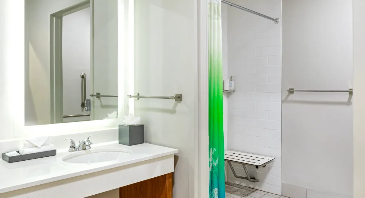 Accessible bathroom with a sink, mirror, tissue box, grab bars, and a shower with a green and blue curtain and a fold-down seat.