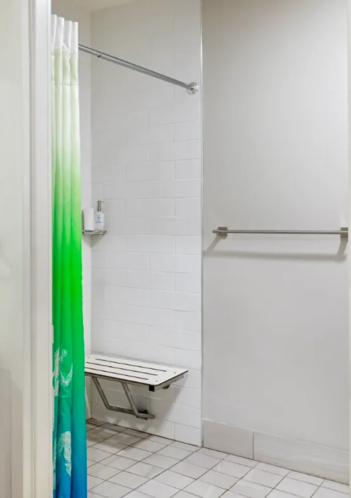 Accessible bathroom with a sink, mirror, tissue box, grab bars, and a shower with a green and blue curtain and a fold-down seat.