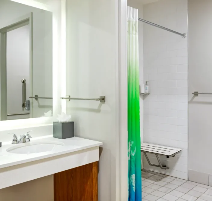 Accessible bathroom with a sink, mirror, tissue box, grab bars, and a shower with a green and blue curtain and a fold-down seat.