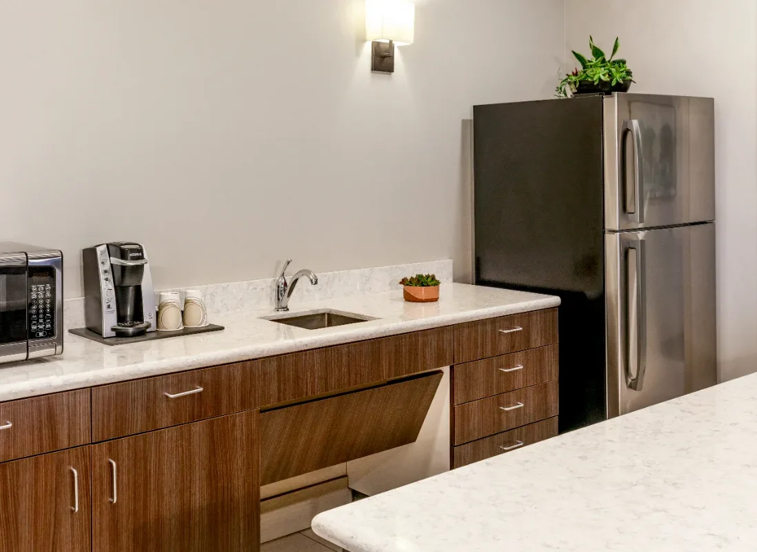 Modern kitchen with brown cabinets, a sink, a silver fridge, a microwave, a coffee maker, and a potted plant on the counter.