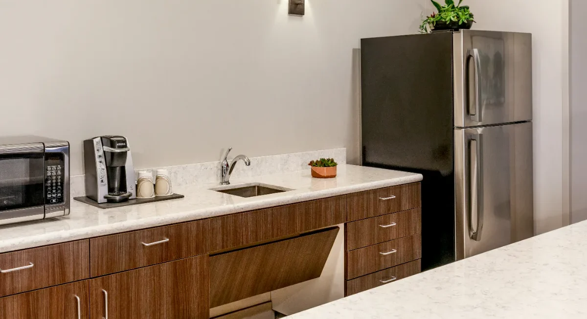 Modern kitchen with brown cabinets, a sink, a silver fridge, a microwave, a coffee maker, and a potted plant on the counter.