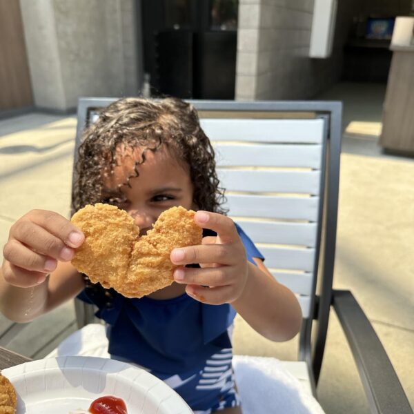 A young child holds up a chicken nugget at an outdoor table, with fries and ketchup on a plate in front of them.