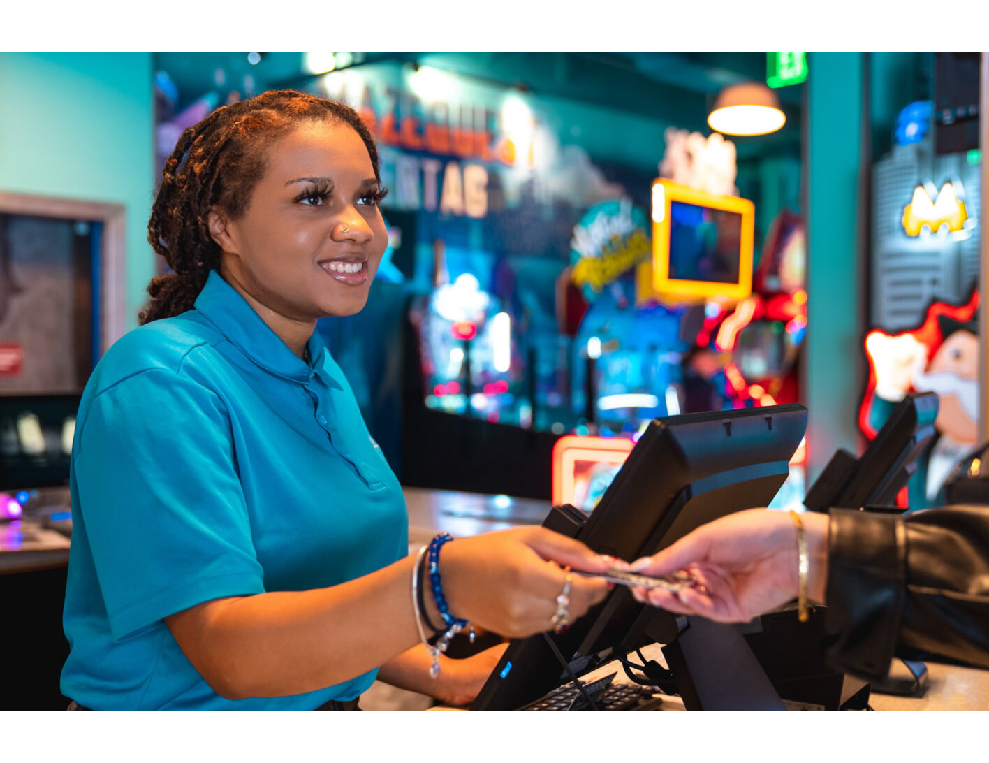 Smiling woman in a blue shirt hands change to a customer at an arcade or entertainment center, with colorful games visible in the background.
