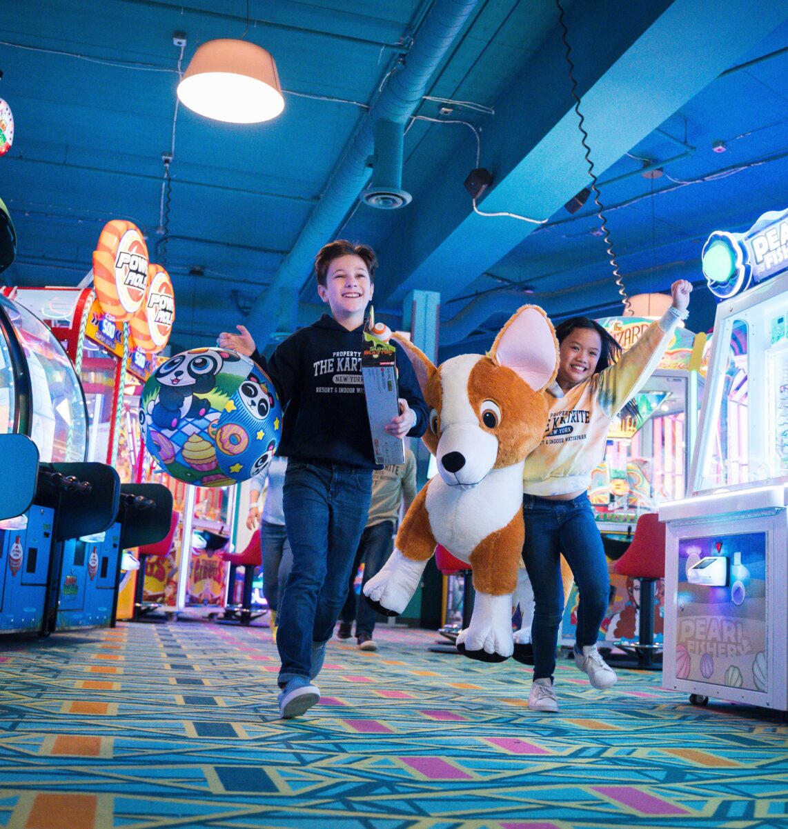 Two kids happily walk in an arcade, carrying large prizes: a stuffed corgi and a colorful ball. Bright lights and games surround them.