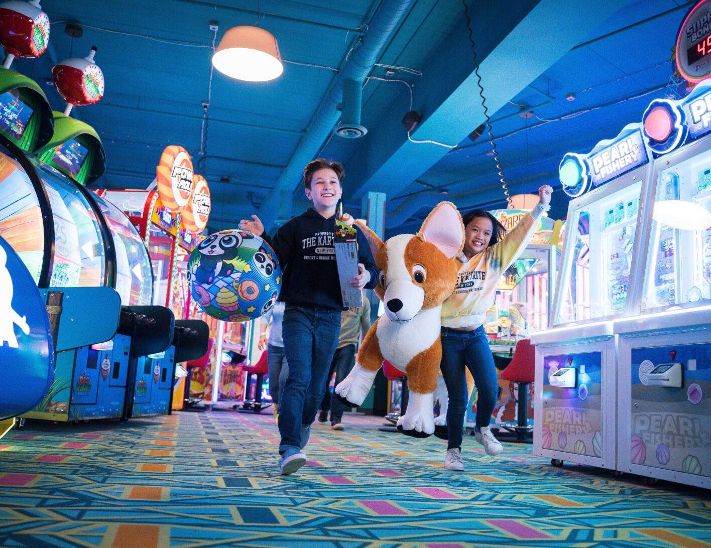 Two kids happily walk in an arcade, carrying large prizes: a stuffed corgi and a colorful ball. Bright lights and games surround them.