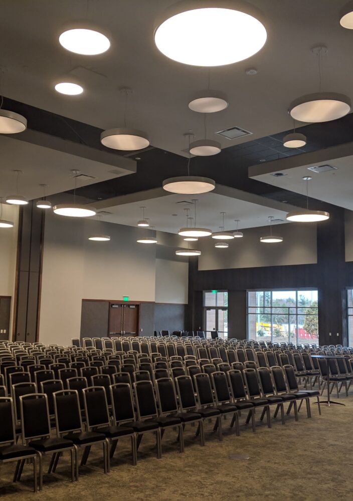 Rows of empty chairs arranged in a large, modern conference room with circular ceiling lights and windows letting in daylight.