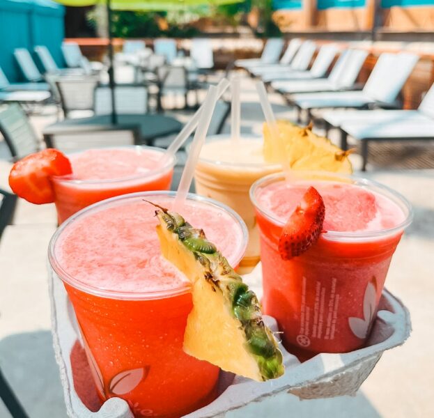 Three bright red smoothies garnished with pineapple and strawberry slices in a drink carrier by lounge chairs at a sunny outdoor pool.