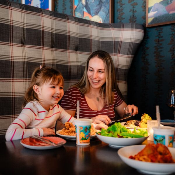A woman and a young girl smile and eat together in a booth, with plates of food and drinks on the table.