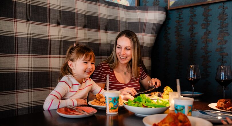 A woman and a young girl smile and eat together in a booth, with plates of food and drinks on the table.