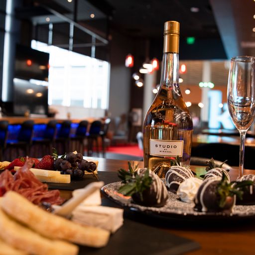 A bottle of white wine, a filled wine glass, and plates of cheese, charcuterie, and chocolate-covered strawberries on a restaurant table.