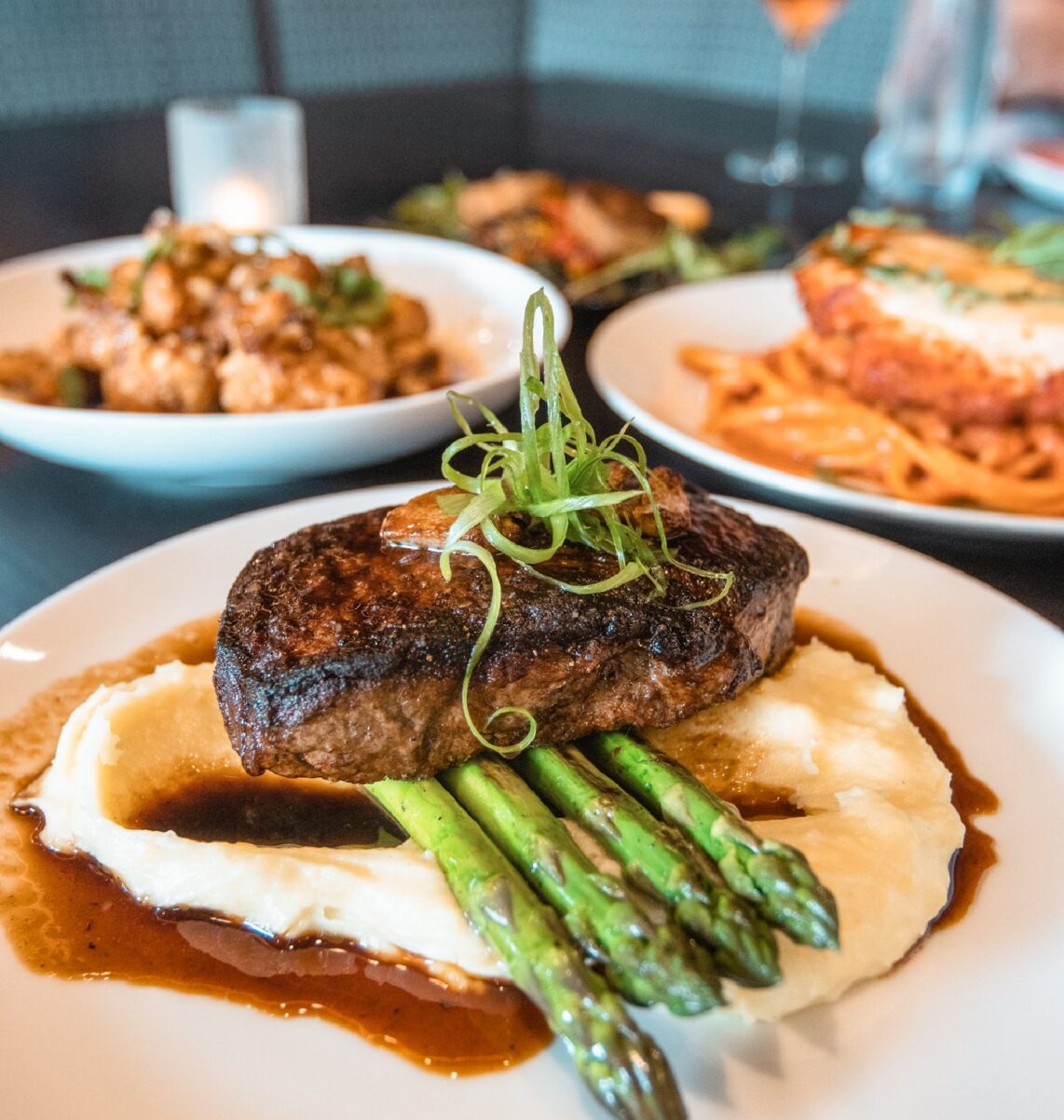 A seared steak on mashed potatoes with asparagus and sauce, garnished with green onions. Other plated dishes are blurred in the background.