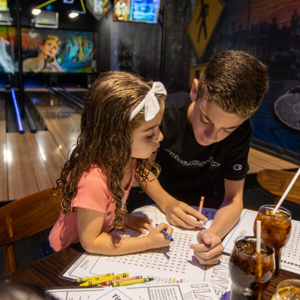 Two kids sit at a table in a bowling alley, working together on a puzzle with crayons. Drinks and menus are on the table beside them.